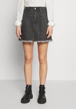 Kvinde iført en sort denim mininederdel med frynset kant og frontslids, kombineret med sorte ankelstøvler med spænde og en hvid top med lange ærmer.