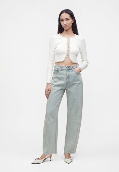 Femme aux longs cheveux foncés portant un cardigan court blanc, un jean large taille haute bleu clair et des chaussures blanches à petit talon.