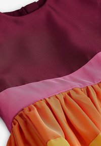 Farbblöckiges Kleid mit einem kastanienbraunen Oberteil, einer rosa Taille und einem orangen, schwingenden Rock mit weicher, fließender Textur.