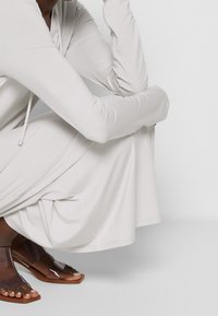 Ljusgrå långärmad klänning med huva och justerbar dragsko. Modell bär genomskinliga bruna sandaler, vilket framhäver plaggets släta textur.