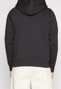 Sudadera con capucha negra de acabado texturizado, con dobladillo plano y puños acanalados, llevada sobre pantalones de color claro.