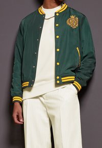 Giacca bomber satinata verde con accenti e bottoni gialli, caratterizzata da un emblema decorativo sul petto e polsini a coste.