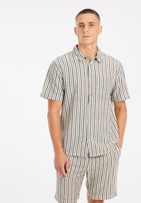 Camisa de manga corta con botones de tela de rayas en beige y marrón, que incluye un bolsillo en el pecho y un corte relajado, combinada con pantalones cortos a juego.