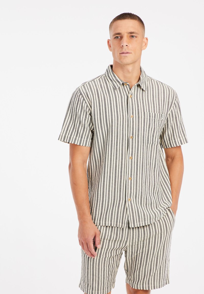 Camisa de manga corta con botones de tela de rayas en beige y marrón, que incluye un bolsillo en el pecho y un corte relajado, combinada con pantalones cortos a juego.