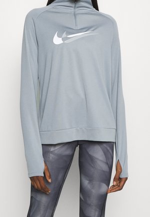 Osoba nosząca jasnoszary, zapinany na zamek, długi rękaw z białym podwójnym logo Nike i ciemne legginsy w wzory.
