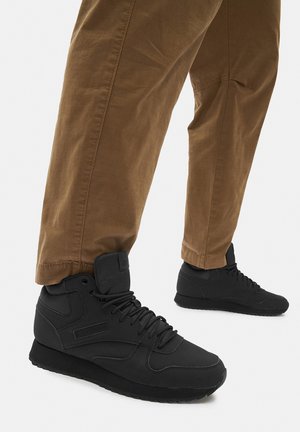 Czarne trampki za kostkę o gładkiej fakturze, ze sznurówkami i gumowymi podeszwami. Noszone z brązowymi, podwiniętymi spodniami o prostych nogawkach.