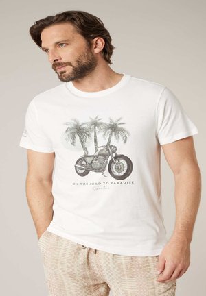 Homme barbu portant un t-shirt blanc avec un motif de moto et de palmiers accompagné du texte « Sur la route du paradis », associé à un pantalon beige à motifs.
