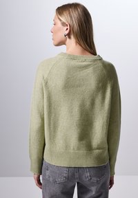 Ljusgrön tröja med mjuk textur, ribbad halsringning och nederkant, med en avslappnad passform och sänkta axelsömmar. Kombinerad med grå jeans.