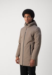 Chaqueta acolchada y con capucha de color marrón, con cremallera frontal y bolsillos laterales; cuenta con puños elásticos y un logo en la manga.