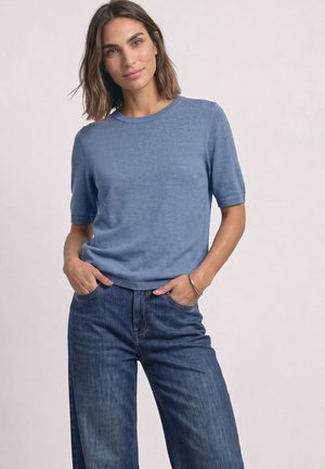 Femme aux cheveux mi-longs, portant un pull à manches courtes bleu clair et un jean bleu foncé, debout les mains dans les poches devant un fond uni.