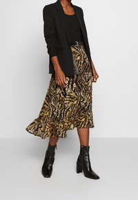 Schwarze Blazer über einem schwarzen Tank-Top, kombiniert mit einem braun-gelben gemusterten Midi-Rock mit Rüsche am Saum und schwarzen Blockabsatz-Stiefeletten.