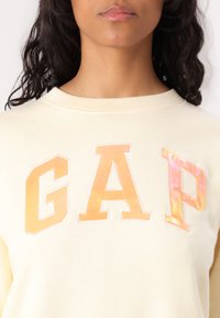 Kremfarget hettegenser med rund hals og myk tekstur, med en blank oransje "GAP" logo i fete bokstaver over fronten.