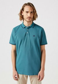 Teal pikétröja med mjuk textur, treknapplås, korta ärmar och kontrasterande marinblå krage och ärmdetaljer.