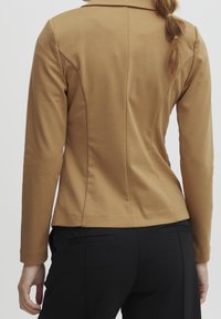 Chaqueta marrón ajustada con mangas largas, costuras estructuradas y un cuello clásico, combinada con pantalones negros que tienen bolsillos laterales.