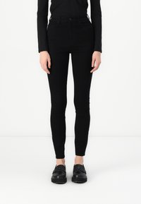 Magasított derekú fekete skinny farmernadrág sima textúrával, elülső zsebekkel és gombos zárással. Fekete slip-on cipővel párosítva.