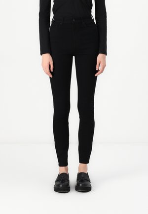 Hoge taille zwarte skinny jeans met een gladde textuur, voorzien van zakken aan de voorkant en een knoopsluiting. Gecombineerd met zwarte instappers.