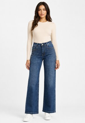 Woman wearing cream ribbed long-sleeve top, high-waisted wide-leg blue jeans with small white dots, and white sneakers, standing against white background.