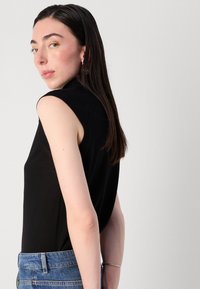 Mladá žena s dlouhými tmavými vlasy, oblečená v černém bezrukávovém topu a modrých džínách, se dívá přes rameno na jednoduchém šedém pozadí.