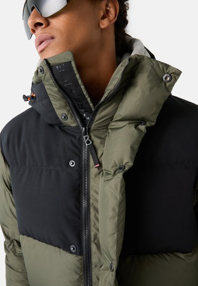 Padded jacket in olive green and black, featuring a zip closure, snap buttons, high collar, and logo detailing along the zipper.
