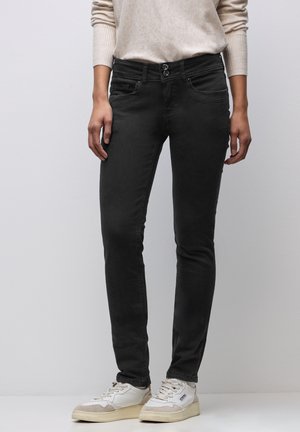 Schwarze Skinny-Jeans mit glatter Textur, ausgestattet mit einem Zwei-Knopf-Verschluss, Fronttaschen und kombiniert mit weißen und beigen Turnschuhen.