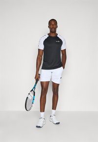 T-shirt de sport noir et blanc avec des manches raglan, associé à un short blanc. Tenant une raquette de tennis, portant des chaussettes et des chaussures blanches.