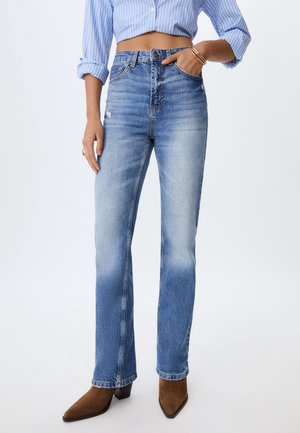 Vrouw draagt lichtblauwe jeans met hoge taille en rechte pijpen, blauw gestreept overhemd met opgerolde mouwen en bruine enkellaarzen met puntige neus.