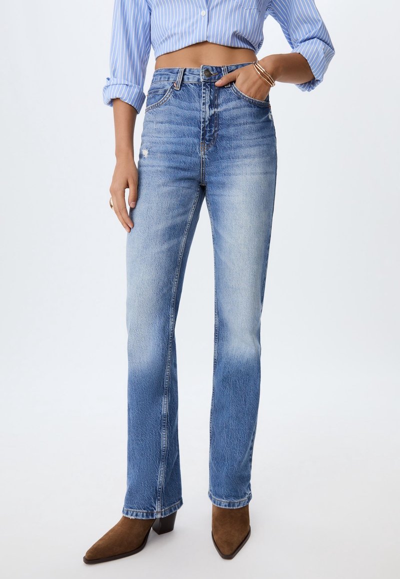 Femme portant un jean bleu clair taille haute à jambes droites, une chemise bleue rayée avec les manches retroussées et des bottines marron à bout pointu.