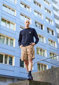 Cămașă albastră cu maneci lungi și buzunar, pantaloni scurți maro cu model, pantofi sport negri și șosete gri. Fundalul are o clădire albastră cu accente galbene.