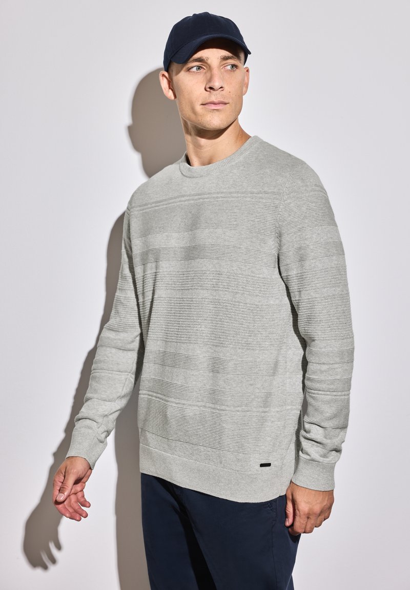 Pull en maille texturée gris clair avec des rayures horizontales, col rond et poignets côtelés, associé à un pantalon bleu marine et une casquette bleu marine.