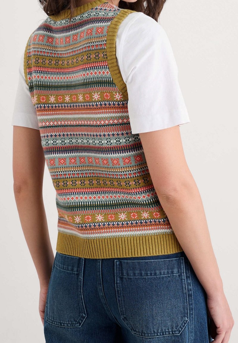 Personne portant un gilet tricoté coloré avec des motifs géométriques par-dessus une chemise blanche à manches courtes et un jean bleu, vue de dos.