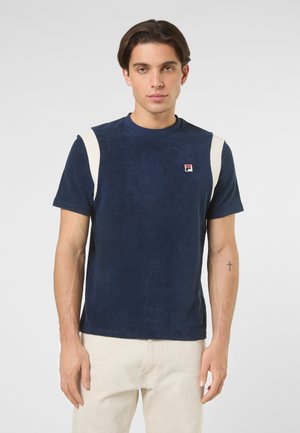 Junger Mann mit kurzem dunklem Haar, der ein marineblau-weißes kurzärmeliges Hemd und beige Hosen trägt und vor einem schlichten Hintergrund steht.