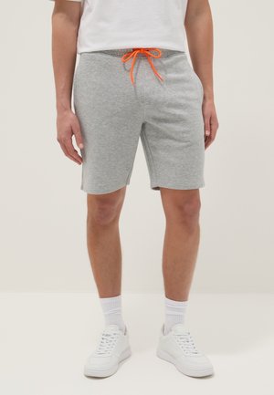 Man wearing light gray knee-length shorts with orange drawstring, white socks, white sneakers, and a white t-shirt, standing against plain background.