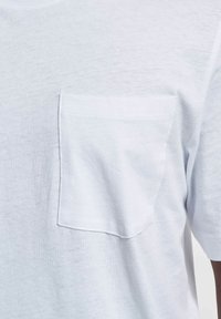 T-shirt bianco in cotone con maniche corte, dotato di una tasca sul petto lato sinistro. Il tessuto ha una finitura morbida e strutturata.