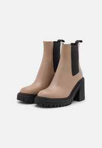 Bottines en cuir beige avec des côtés élastiques noirs, semelle en caoutchouc noir épaisse et talon carré. Design minimaliste avec une texture lisse.
