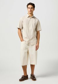 Camicetta beige a righe con maniche corte e bottoni, abbinata a pantaloni corti color avorio fino al ginocchio e scarpe da barca marrone scuro con dettagli in pizzo.