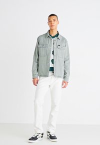 Camisa de denim gris claro con dos bolsillos frontales, llevada abierta sobre un suéter de patrón verde y blanco, combinada con jeans blancos y zapatillas negras.