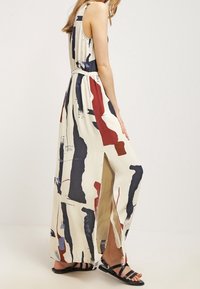 Robe maxi avec un fond crème, ornée de motifs abstraits bleu marine, rouge et noir. Design sans manches avec une haute fente et un détail de taille nouée.