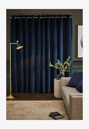Dunkelblaue Vorhänge hängen hinter einem beigen Sofa mit blauen Kissen, einem runden Beistelltisch mit einer Vase mit Grünpflanzen und einer goldenen Stehlampe.