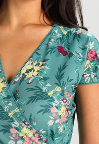 Květinové volánové tričko v tónech smaragdově modré, se výstřihem do V a krátkými rukávy. Tkanina má vzor s žlutými, růžovými a červenými květy na zeleném pozadí.