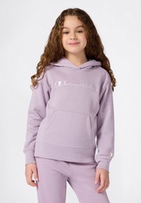 Niña pequeña con cabello rizado, vistiendo una sudadera de Champion color lavanda y pantalones a juego, de pie frente a un fondo blanco liso.