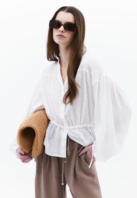 Top bianco leggero con bottoni sul davanti, maniche a sbuffo e vita arricciata. La modella tiene in mano una borsa intrecciata color cammello. Sono visibili pantaloni marroni.