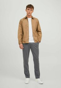 Beige Hemdjacke über einem weißen T-Shirt, kombiniert mit grauen Sweatpants. Model trägt weiße Sneakers. Zwei Brusttaschen auf der Jacke.