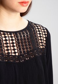 Schwarze Bluse mit einem strukturierten, gehäkelten Spitzeneinsatz und geometrischen Mustern. Weicher Stoff, runder Ausschnitt und fließende Passform.