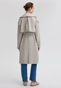 Heledad beige mantli, millel on lühendatud seljatagune, pikad varrukad ja vöökoht, kombineeritud siniste teksade ja beežide kingadega, vaadatud tagant.