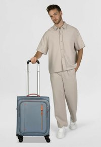 Valigia rigida blu con texture liscia, dettagli arancioni e manico retrattile, dotata di quattro ruote e tasca frontale con cerniera.