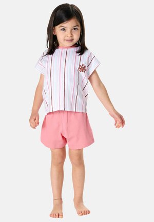 Girl standing barefoot wearing a striped short-sleeve shirt with a floral detail and pink shorts against a plain background.