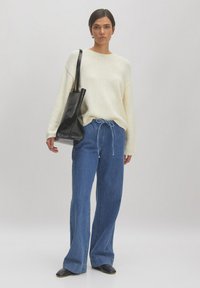 Cremfarbener Strickpullover, lockere Passform, gerippte Bündchen; hochgeschnittene blaue Jeans, weit geschnitten; schwarze Tragetasche; schwarze Lederschuhe, glatte Oberfläche.