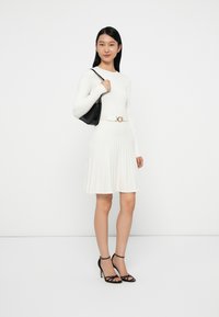 Abito bianco a coste con maniche lunghe, corpetto aderente e gonna svasata. Accentuato da una borsa nera e tacchi neri con cinturini.