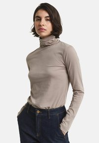 Frau mit kurzen dunklen Haaren, die einen beigen Rollkragenpullover und dunkelblaue Jeans trägt und mit den Händen in den Taschen steht, vor einem weißen Hintergrund.