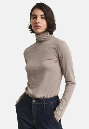Frau mit kurzen dunklen Haaren, die einen beigen Rollkragenpullover und dunkelblaue Jeans trägt und mit den Händen in den Taschen steht, vor einem weißen Hintergrund.